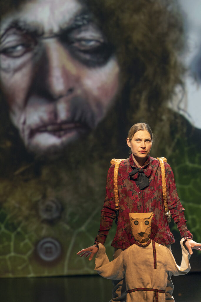 Valokuvan taka-alalla on piirroskuva karvaisesta jättiläisestä ja etualalla on näyttelijä ja Antti Puuhaara -nukke.