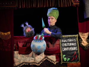 Kuvassa Raxu Taskunen -nukke ja esiintyjä Simo Heiskanen, joka pitää kädessään chiliä hämmentynyt ilme kasvoillaan.