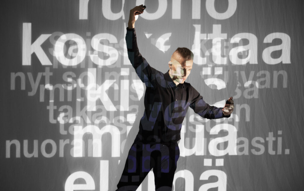 Kuvassa on esiintyjä Simo Heiskanen, joka katsoo alaspäin ojennettua kättään. Esiintyjän taakse on projisoituna erilaisia runonpätkiä.
