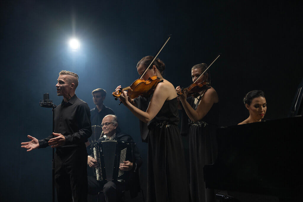 Etualalla pianista ja kaksi viulunsoittajaa, heidän takaansa näkyy laulaja, joka laulaa mikrofoniin. Laulajan takana näkyy harmonikan soittaja. Kuva on otettu sivulta päin.