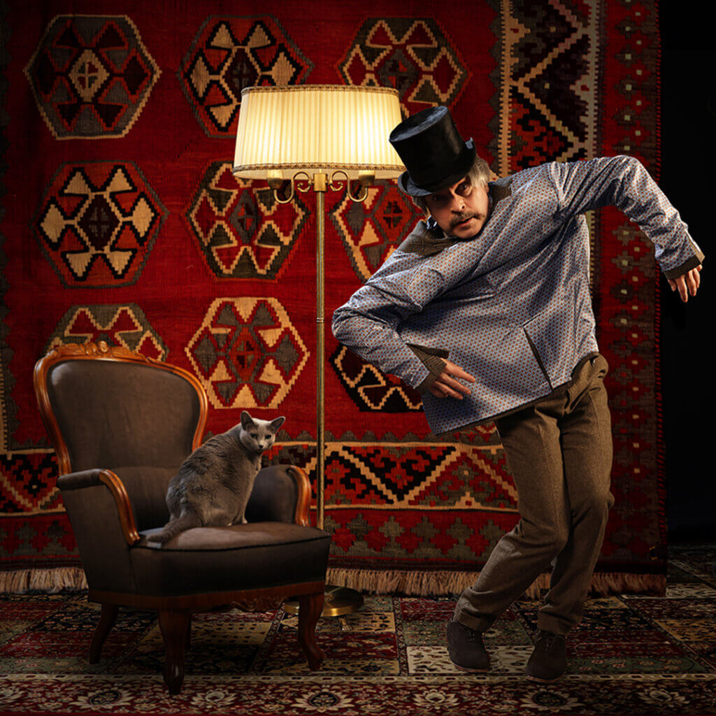 Vasemmalla harmaa kissa istuu nojatuolissa, oikealla puolella henkilö on pukeutunut silinterihattuun ja esittää variksen pelätintä, heidän keskellään on jalkalamppu.