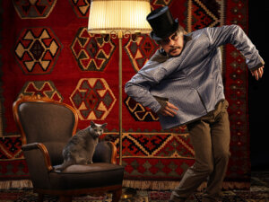 Vasemmalla harmaa kissa istuu nojatuolissa, oikealla puolella henkilö on pukeutunut silinterihattuun ja esittää variksen pelätintä, heidän keskellään on jalkalamppu.