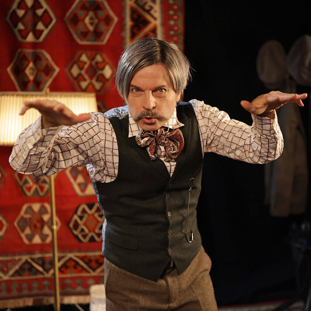 Henkilö, jolla on harmaat hiukset ja viikset, on nostanut molemmat kädet ilmaan ja näyttää, kun hän tekisi loitsuja.